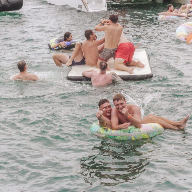 A group of people enjoy swimming and lounging on inflatables and a floating platform in a lake. Two men smile at the camera from a colorful inner tube in the foreground. The scene is lively and playful.