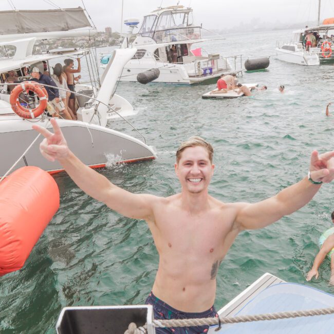 A smiling shirtless man stands on a boat, flashing peace signs with both hands. Several people swim and relax on floaties in the water, with multiple boats anchored nearby in a lively, festive atmosphere.