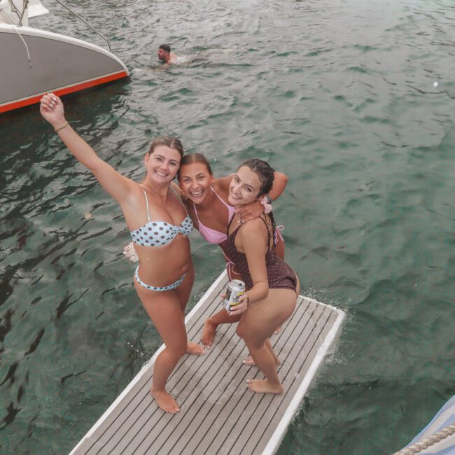Three smiling women in swimsuits pose together on a boat's swim platform above the water, with one raising her arm. A swimmer is in the water nearby and parts of a boat are visible. The mood is cheerful and festive.
