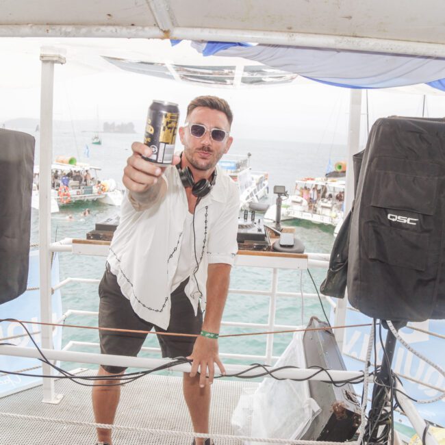A man wearing sunglasses and headphones holds up a can while standing between speakers on a boat, with water, other boats, and people visible in the background.