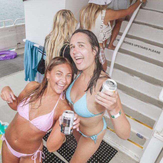Two women in bikinis smile and hold canned drinks on a boat, appearing happy and wet as if they’ve been swimming. Other people are seen in the background near a staircase. The scene is bright and festive.