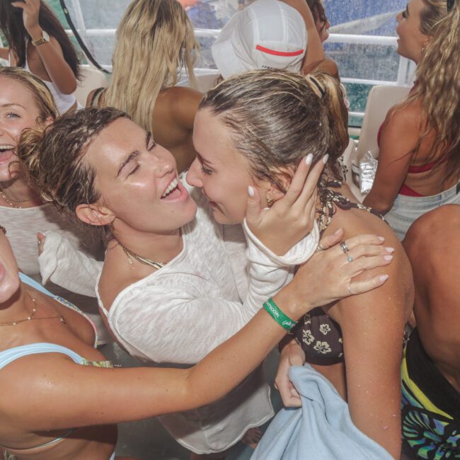 A group of young people in swimsuits laugh, smile, and hug each other on a crowded boat, with water splashing around them during a lively, sunny party.