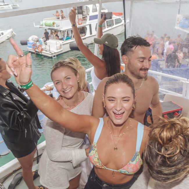 A group of young adults smile and dance on a boat during a lively party, with several boats and people on the water in the background. The atmosphere is joyful and festive.
