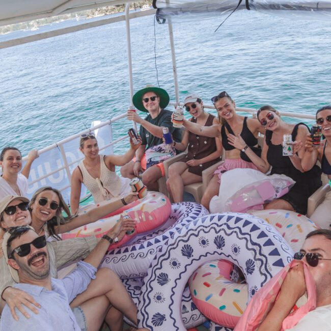 A group of people smile and pose for a photo on a boat decorated with colorful pool floats, holding drinks and phones, with blue water and another boat visible in the background.