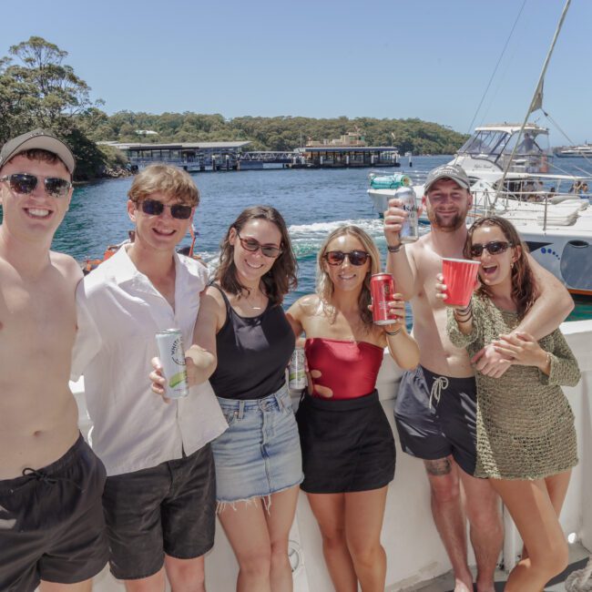 Six young adults stand on a boat in swimwear and summer clothes, smiling and holding drinks, with a marina, sailboats, and trees in the background under a bright blue sky.