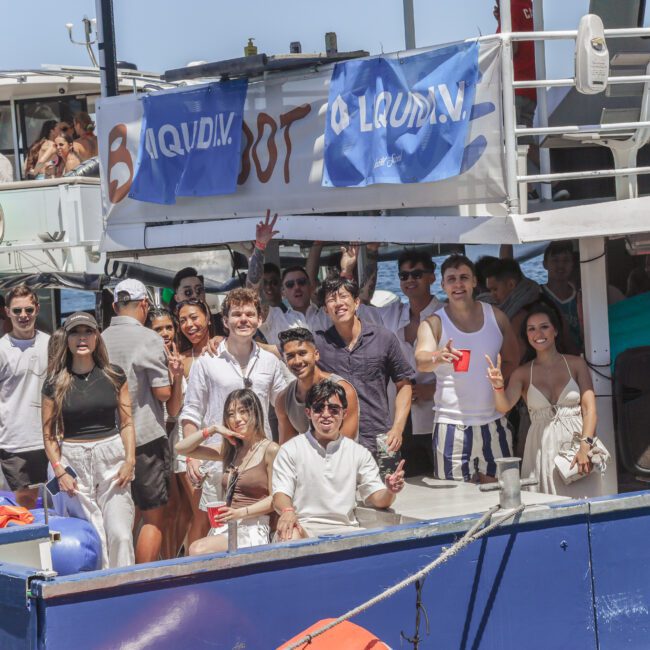 A group of people smiling and posing for a photo on the deck of a boat during a sunny day, with blue banners and party decorations visible in the background.