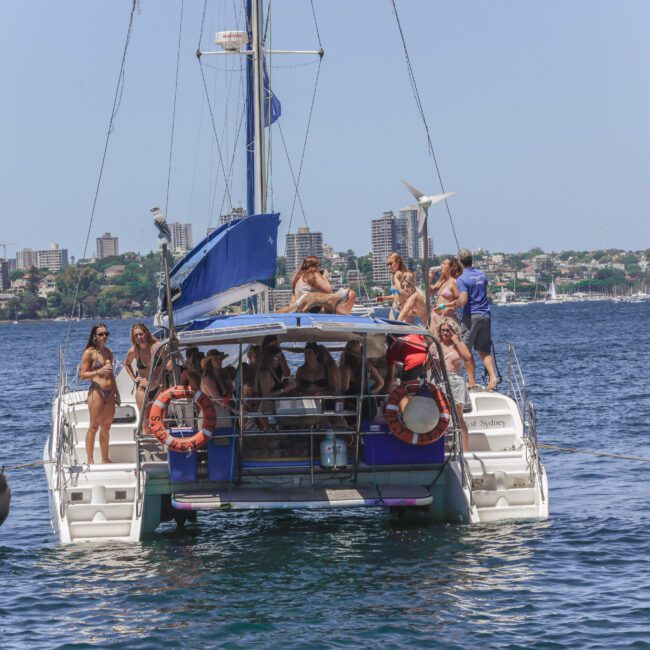 A group of people in swimsuits are gathered on the deck of a catamaran anchored on the water, enjoying a sunny day with a city skyline and greenery in the background.