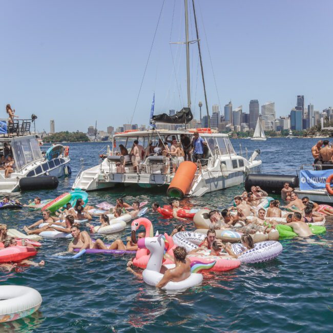 A lively group of people relax on colorful inflatables in the water near several anchored boats, with a city skyline in the background under a clear blue sky.