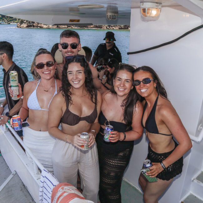A group of young adults in swimwear smile and pose on the deck of a boat, holding drinks, with water and a coastline visible in the background on a sunny day.