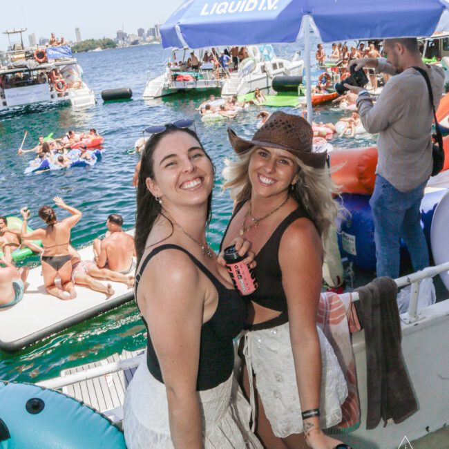 Two smiling women pose on a boat holding drinks at a lively yacht party with many people swimming, lounging on floats, and socializing in the sun. A large umbrella and city skyline are visible in the background.