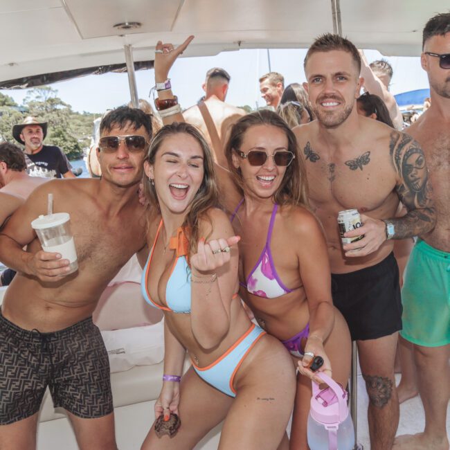 A group of smiling young adults in swimwear pose together on a boat, holding drinks and enjoying a sunny day. Some have sunglasses, and one person holds up a hand in a fun gesture. The atmosphere is lively and festive.