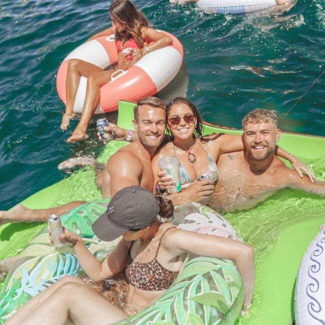 Four people relax on inflatable rafts in clear blue water, smiling and holding canned drinks, while another person floats nearby on a round inflatable. The atmosphere is sunny and joyful.