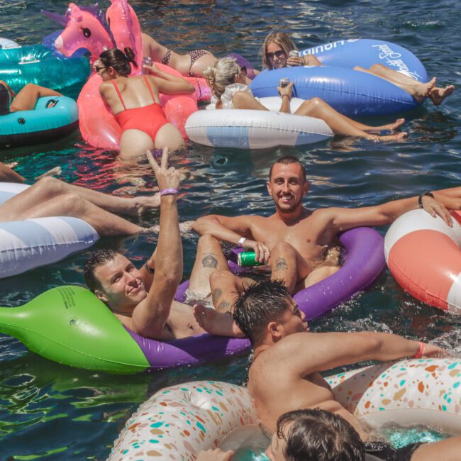 A group of people relax on colorful inflatable pool floats in a crowded, sunlit body of water, smiling, laughing, and enjoying a festive summer atmosphere.