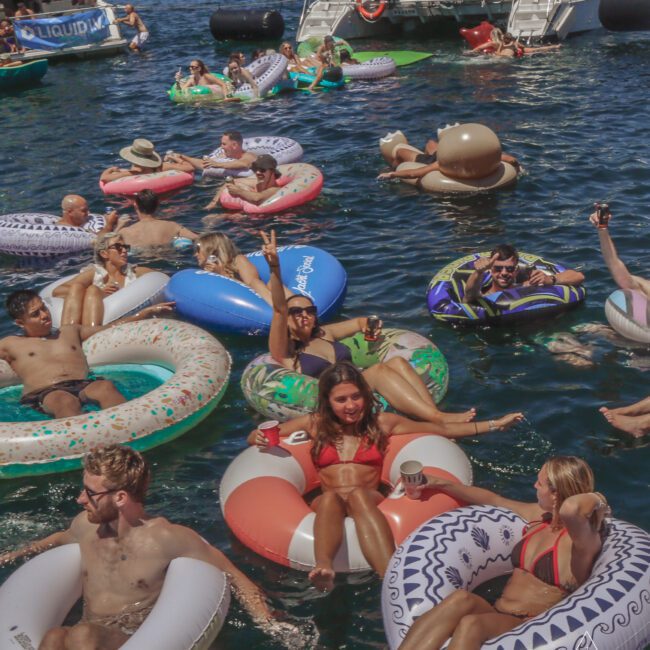 A group of people relax and socialize on colorful inflatable pool floats in the water near docked boats, enjoying a sunny day. Some are holding drinks and smiling, with a festive atmosphere.