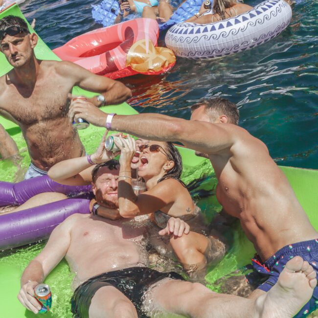 A group of young adults in swimsuits relax and have fun on a floating mat in the water, sharing drinks and laughing, surrounded by inflatable pool floats under bright sunlight.