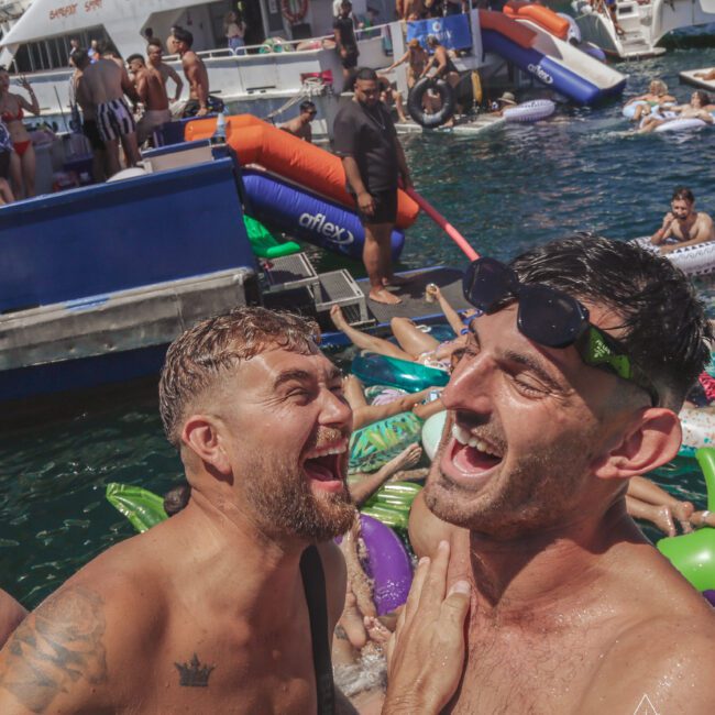 Two men laughing and embracing in the foreground at a lively pool party, surrounded by people on inflatables in the water, with boats and more partygoers visible in the background.