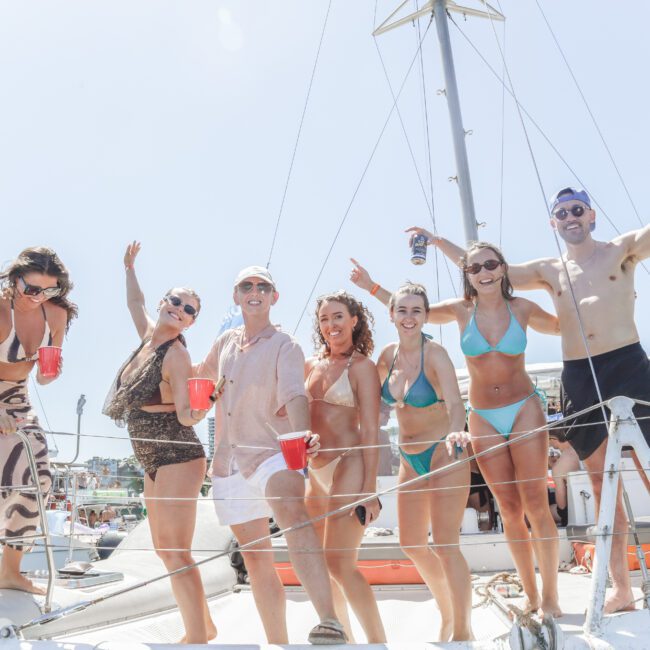 A group of seven people in swimsuits and summer outfits smiling and holding drinks, standing on the deck of a boat under sunny skies. Some have arms raised, celebrating and enjoying a day on the water.