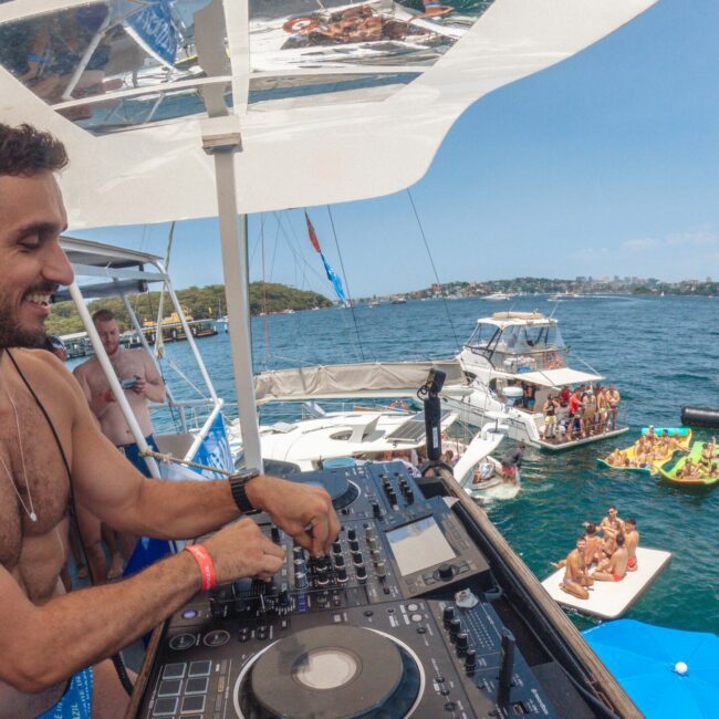 A man in swim trunks and headphones DJs on a boat deck, smiling, with people relaxing and dancing on nearby boats and inflatables on a sunny day over blue water.