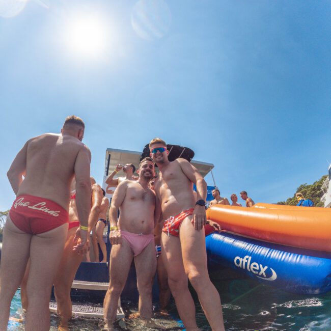 A group of men in swimsuits stand on a floating platform by a boat under the bright sun, with inflatable slides and water toys nearby, enjoying a lively day on the water.
