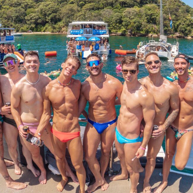 A group of smiling men in colorful swim briefs pose together on a boat deck, with several boats, people swimming, and lush green trees in the background under a sunny blue sky.