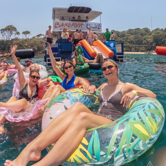 A group of people relax and smile on colorful inflatable tubes in the water near a boat with slides. The sun is shining, and everyone looks happy, enjoying a summer day.