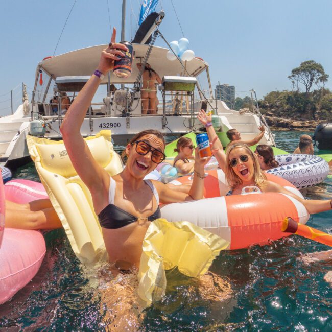 Three women in swimsuits smile and cheer while floating on inflatables in the water near a boat. Others are on the boat, and the scene is festive and sunny, suggesting a fun social gathering.