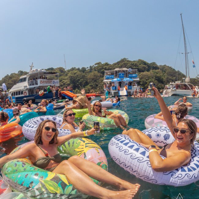 A group of people relax and smile on colorful inflatable floaties in the water near yachts, enjoying a sunny day at a social event. Many hold drinks, and there are forested hills in the background.
