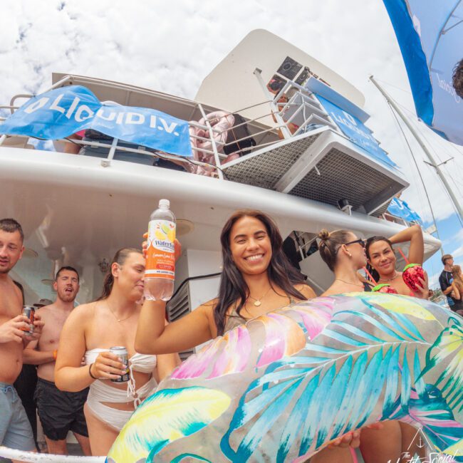 A smiling woman in a bathing suit holds a vitamin drink and a colorful pool float at a lively boat party with other people on deck under a partly cloudy sky.