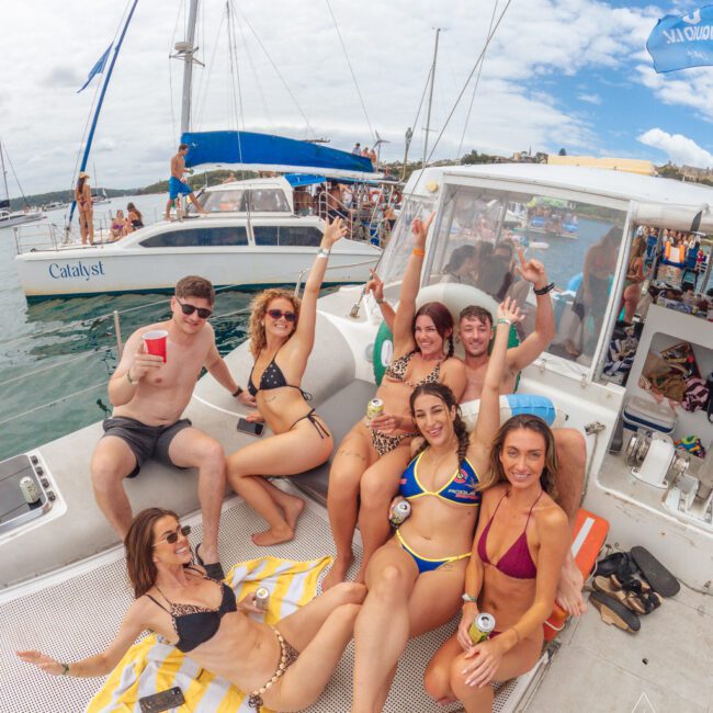 A group of young adults in swimsuits relax and pose, smiling on the deck of a boat at a lively party. Other boats and people are visible in the background, with an Australian flag flying overhead.