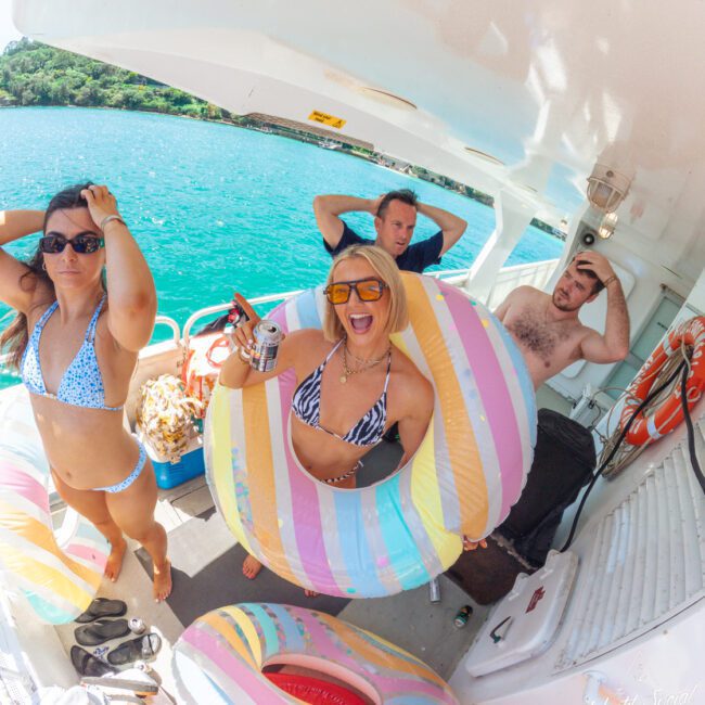 Four people in swimsuits stand on a boat with colorful pool floats, near turquoise water and lush green scenery. One woman in the center smiles holding a drink, while others pose playfully, some with hands on their heads.