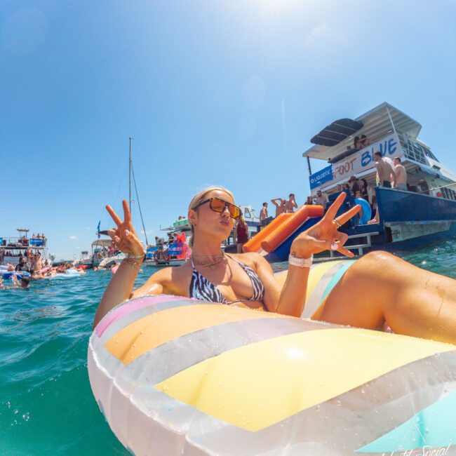 A woman wearing sunglasses and a bikini relaxes on a colorful pool float, flashing peace signs, in clear blue water near a party boat under a sunny sky with other people and boats in the background.