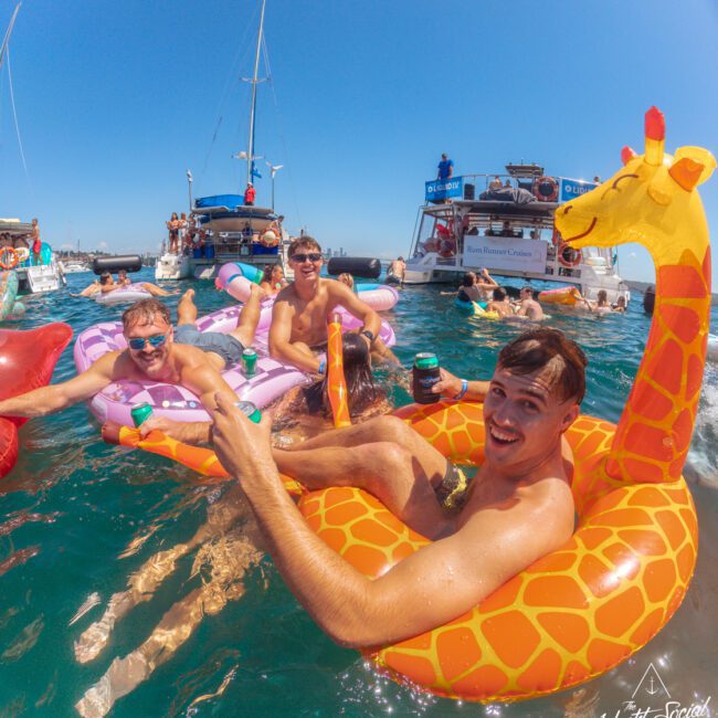 Three young men smile and relax on animal-shaped inflatables, including a giraffe, in clear blue water near anchored boats during a sunny day. Other people and party boats are visible in the background.