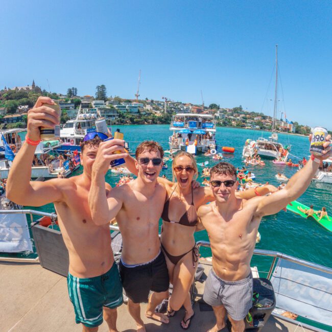 Four young adults in swimwear smile and raise drinks on a dock or boat, with a lively scene of people, boats, and clear blue water in the background on a sunny day.