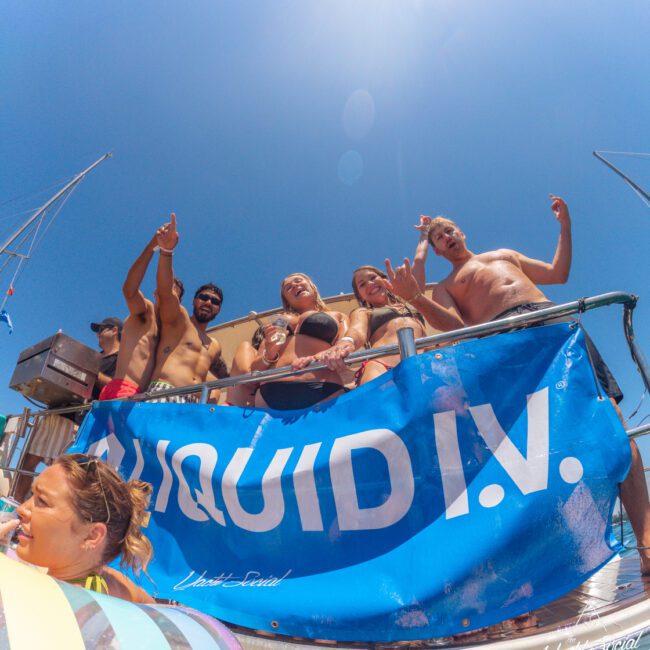 A group of people in swimsuits stand on a boat deck, smiling and making hand gestures behind a blue "LIQUID I.V." banner. The sky is clear and blue, and a pool float is visible in the water below.