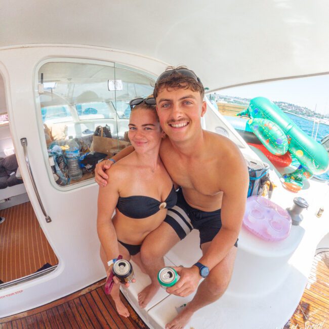 A smiling man and woman in swimsuits sit together on a boat, each holding a can. The boat deck and water with inflatable toys are visible in the background on a sunny day.