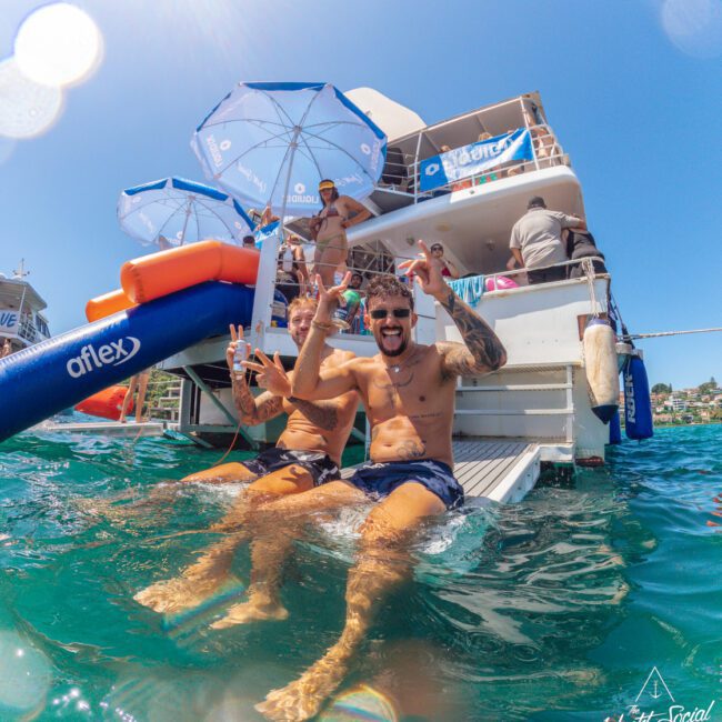 Two shirtless men sit smiling on a waterslide off the back of a yacht, making peace signs. Other people are on the boat under umbrellas. The scene is sunny, with bright blue water and a festive atmosphere.