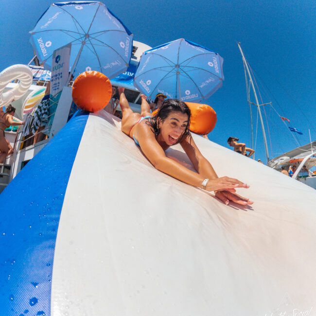 A woman slides face-first down a large inflatable water slide, smiling. Blue umbrellas and people are in the background under a clear sky, with boats visible nearby.