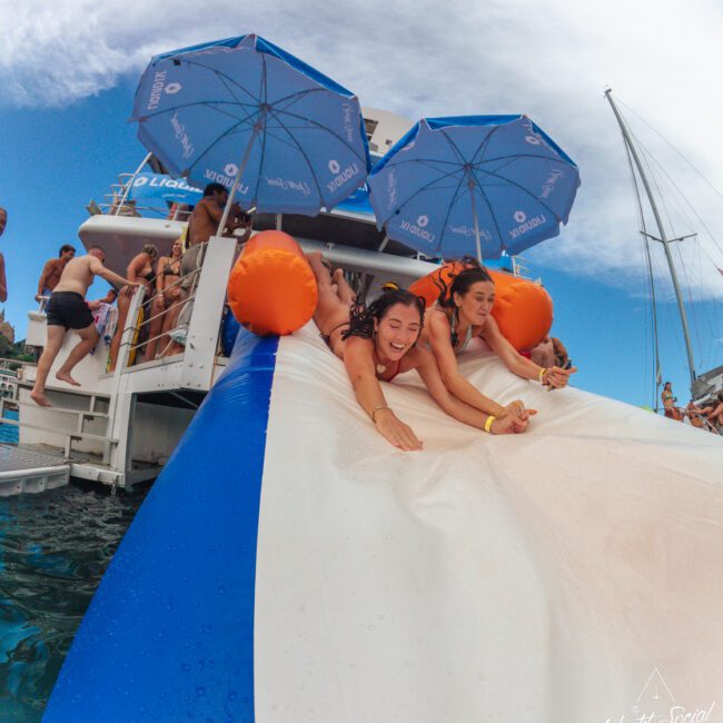 Two women slide down an inflatable water slide from a yacht into the sea, smiling and laughing, while others wait at the top under blue umbrellas. A sailboat and coastline are visible in the background.