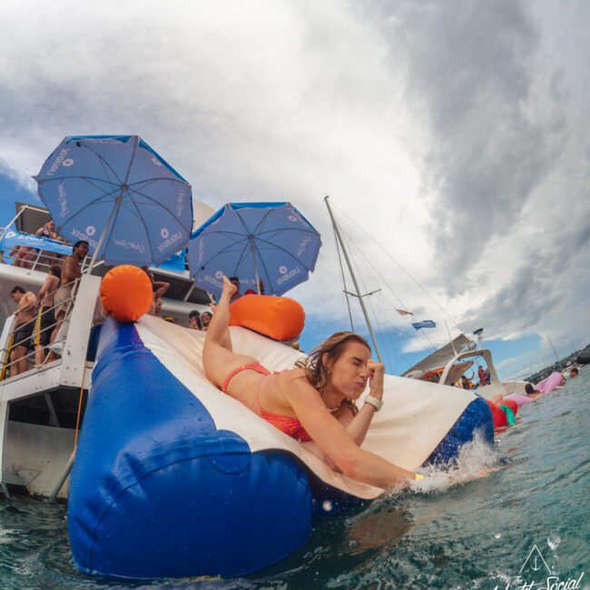 A woman in a red swimsuit slides down an inflatable slide from a boat into the ocean, with people, blue umbrellas, and sailboats in the background under a cloudy sky.
