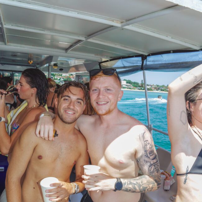 Two smiling men in swim trunks pose together on a crowded boat, each holding a drink. Sunlight shines on them and the blue ocean and coastline are visible in the background.