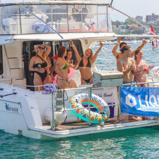 A group of people in swimsuits dance and celebrate on the deck of a white yacht in sunny weather, with water floats and a blue “LIQUID” banner hanging on the side, near a scenic shore.