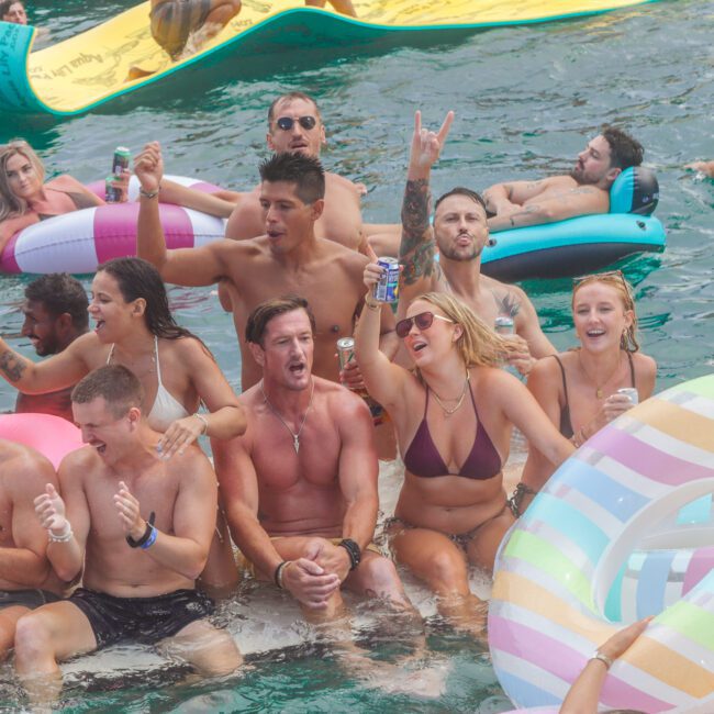 A group of people in swimsuits laugh, cheer, and hold drinks while sitting on inflatables and a floating mat, enjoying a lively party on the water. The mood is festive and energetic.