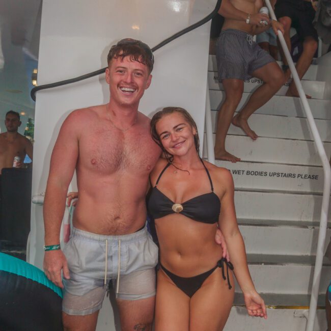 A smiling man in gray swim trunks and a woman in a black bikini pose together on a boat near a staircase, with other people in swimsuits in the background. The atmosphere looks wet and lively.