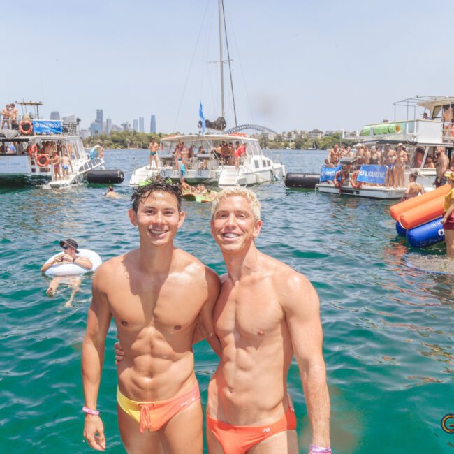 Two men in orange swim briefs smile and pose together in the water at a lively boat party. Behind them, people socialize and relax on boats and inflatables under a sunny sky.