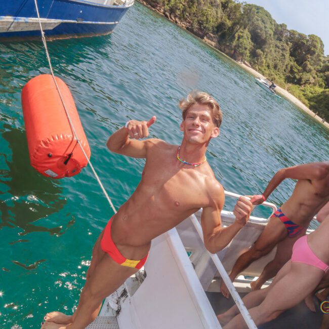 A smiling man in a colorful swim brief stands on a boat, giving two thumbs up. Several other people in swimsuits are gathered on the boat, with clear green water and a tree-lined shore in the background.