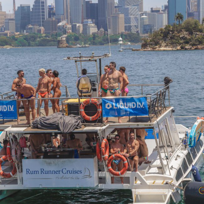 A group of people in swimsuits stands and socializes on a boat labeled "Rum Runner Cruises" on the water, with a city skyline and greenery visible in the background.
