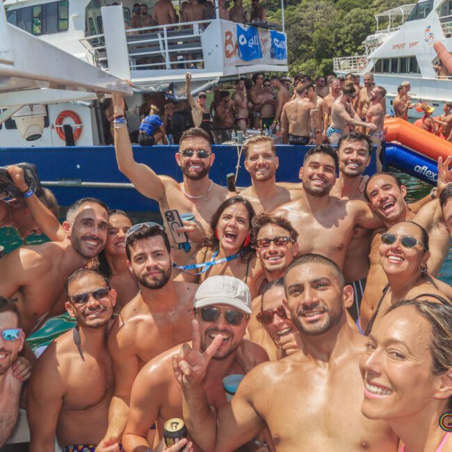 A large group of people in swimwear smile and pose for a selfie on a boat surrounded by water, with more people and boats in the background, enjoying a sunny day at a lively party.