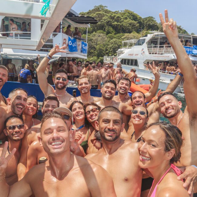 A large group of smiling people in swimwear pose for a selfie on a crowded boat during a sunny day, with more people and boats visible in the background near lush green hills.
