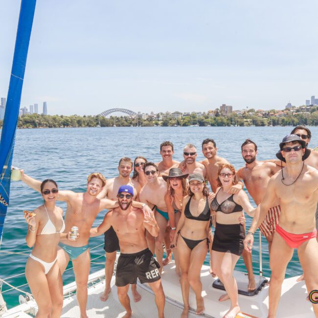 A group of people in swimsuits smiles and poses on a sailboat with the Sydney skyline and Harbour Bridge in the background on a sunny day. The mood is festive and relaxed. The GAYM logo is visible in the corner.