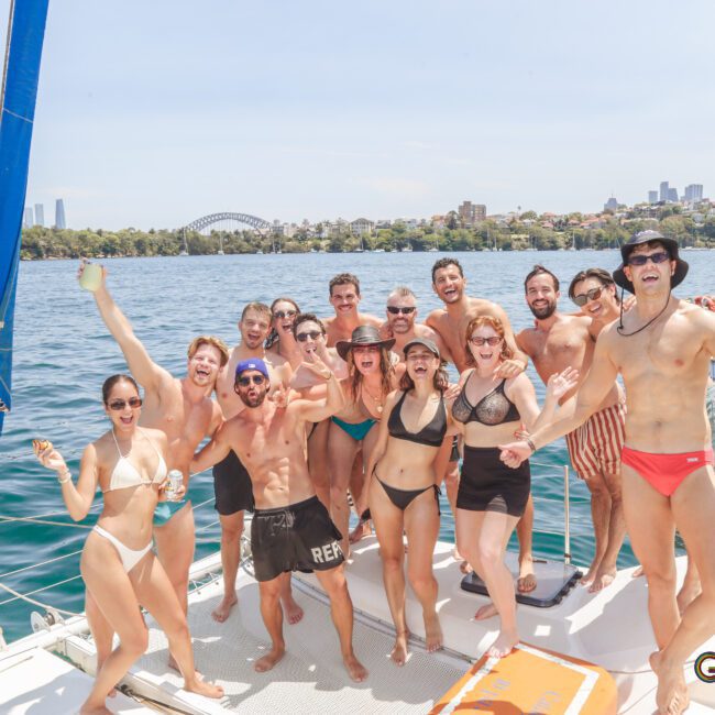 A group of people in swimwear pose and smile on a sailboat with a city skyline and Sydney Harbour Bridge in the background under a sunny sky. The mood is joyful and festive.